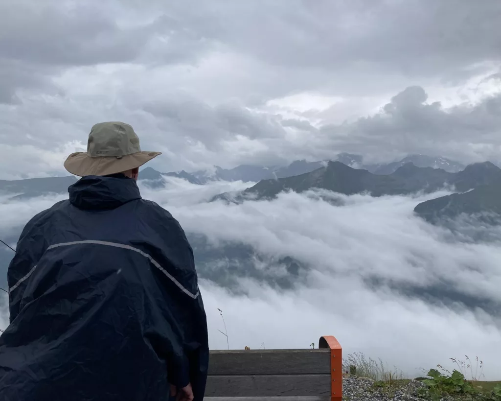 ein Mann mit Regencape und Hut schaut in die Ferne - über den Nebel- und Regenwolken in den Bergen