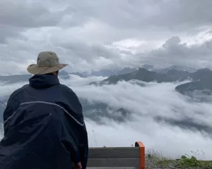 ein Mann mit Regencape und Hut schaut in die Ferne - über den Nebel- und Regenwolken in den Bergen