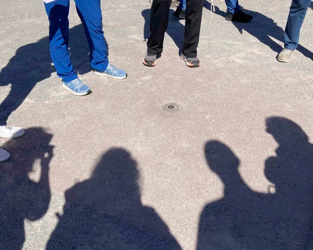 Füße und Schatten einer Menschengruppe, die um eine Messingplatte im Boden im Kreis herumstehen